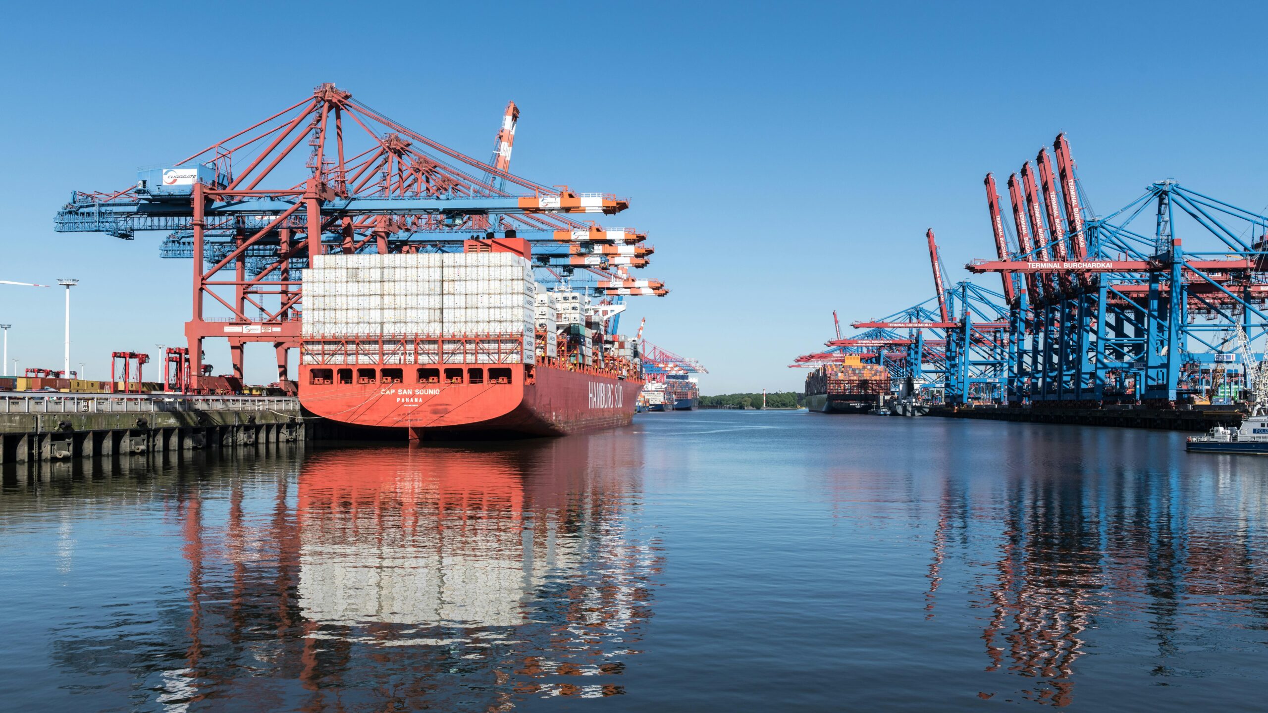 Vibrant container ship docked at Hamburg harbor, reflecting on calm water under a clear blue sky.